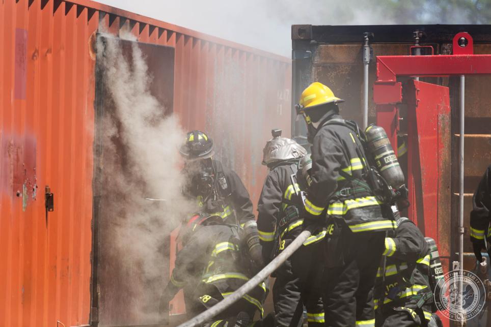 firefighters entering a metal structure with a large hose