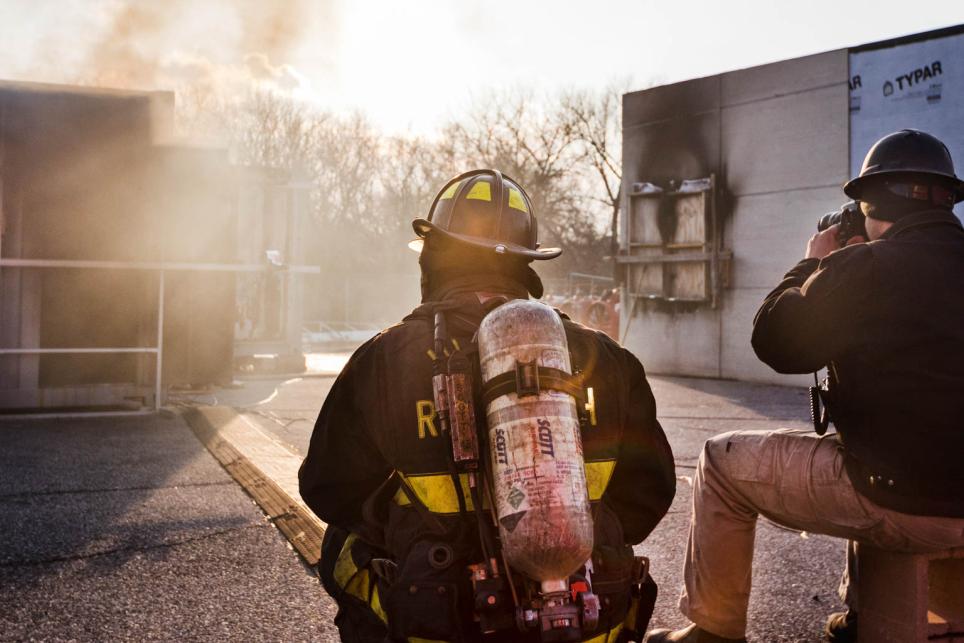 firefighter in full personal protective equipment and research engineer watch smoke coming out of a metal storage container during an experiment.