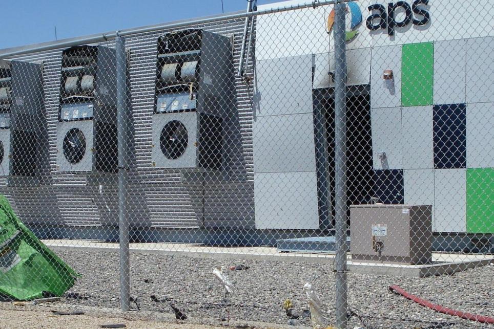 Energy storage system with walls bowed out and broken fence with the door to system leaning on fence after an explosion