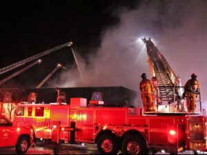 Chula Vista Fire Department crews respond to a warehouse fire. A ladder truck is visible in the foreground, and four extended ladders surround the warehouse, spraying water onto the structure during a near-miss incident.