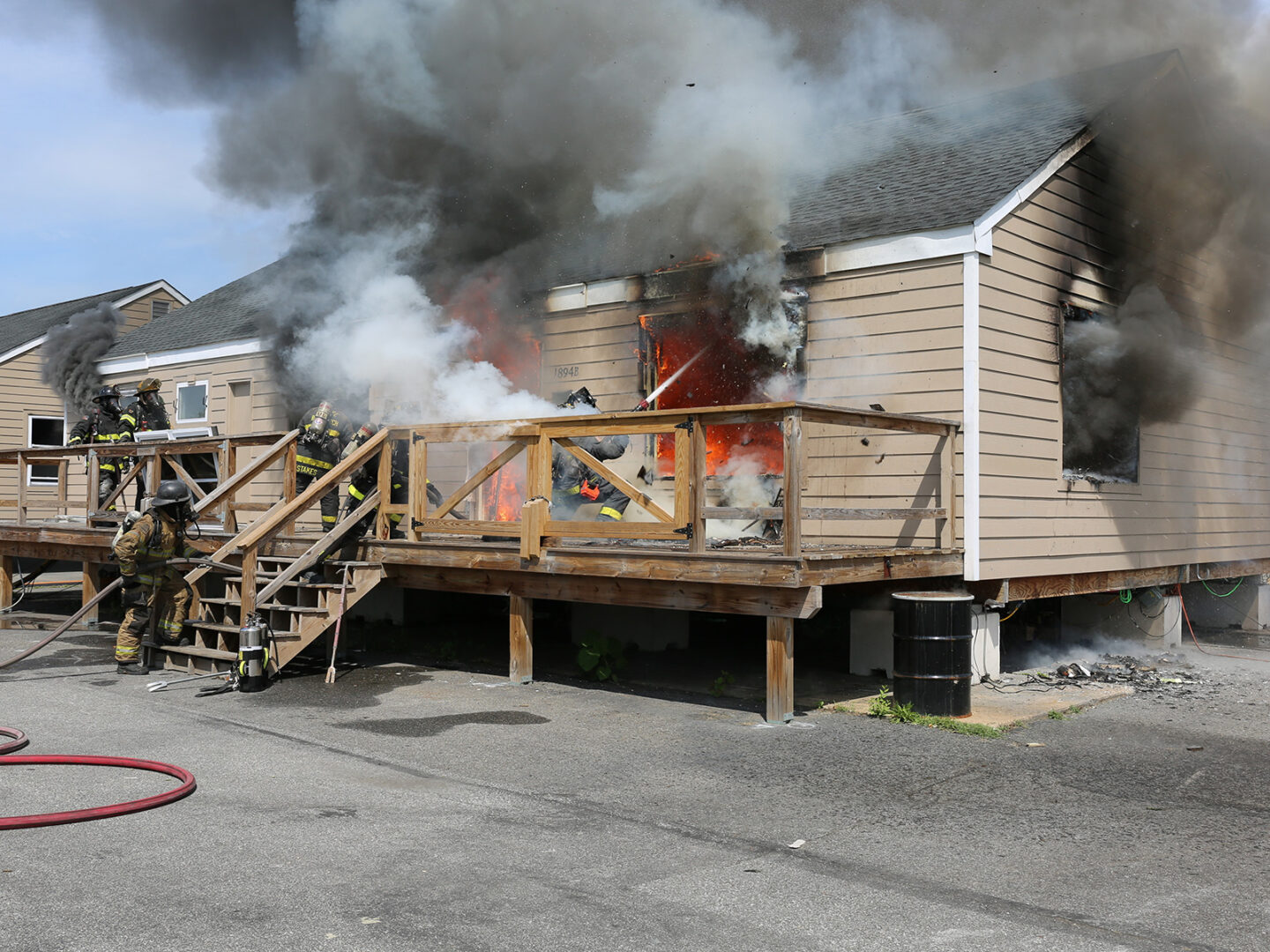 Alt text: Exterior view of a purpose‑built single‑family test structure during an experimental burn. Side A and part of Side D are visible. A failed window on the right side of Side A is centered in the frame, where a firefighter kneels outside, directing a straight stream into the opening. Smoke billows from every window and door as crews respond.