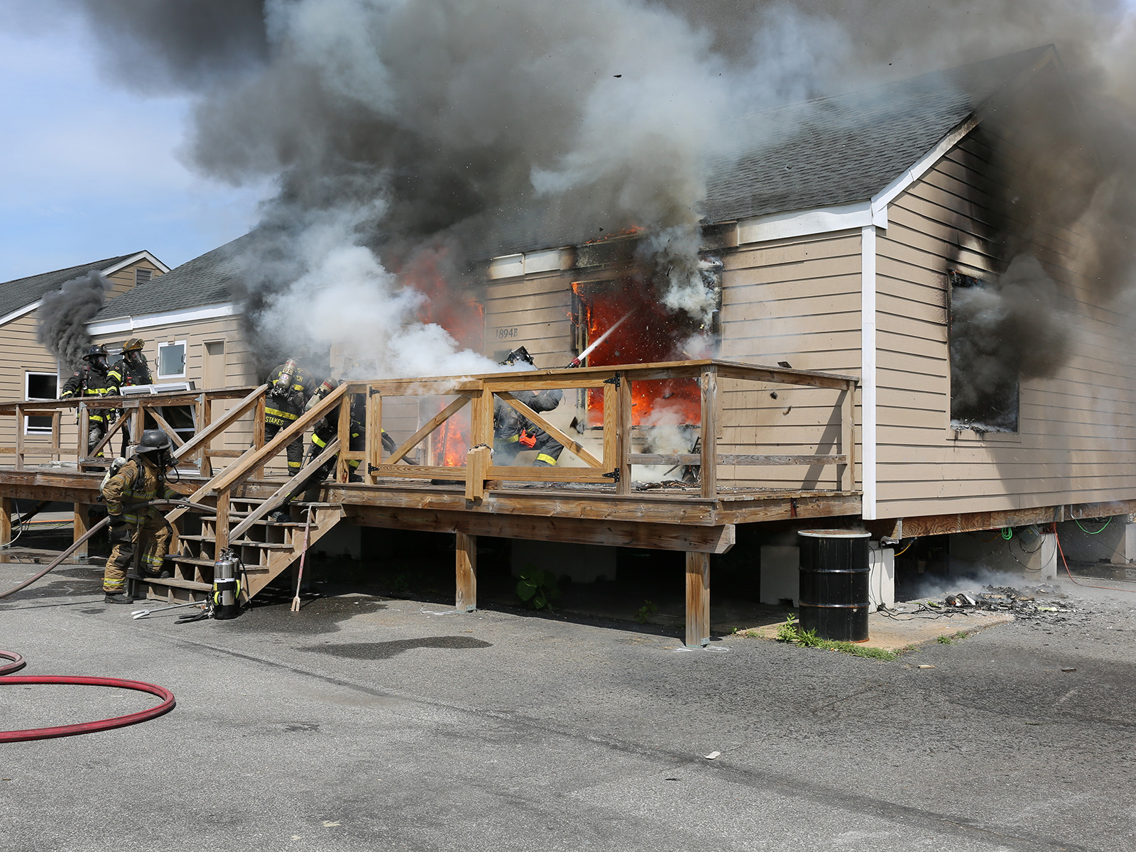 Alt text: Exterior view of a purpose‑built single‑family test structure during an experimental burn. Side A and part of Side D are visible. A failed window on the right side of Side A is centered in the frame, where a firefighter kneels outside, directing a straight stream into the opening. Smoke billows from every window and door as crews respond.