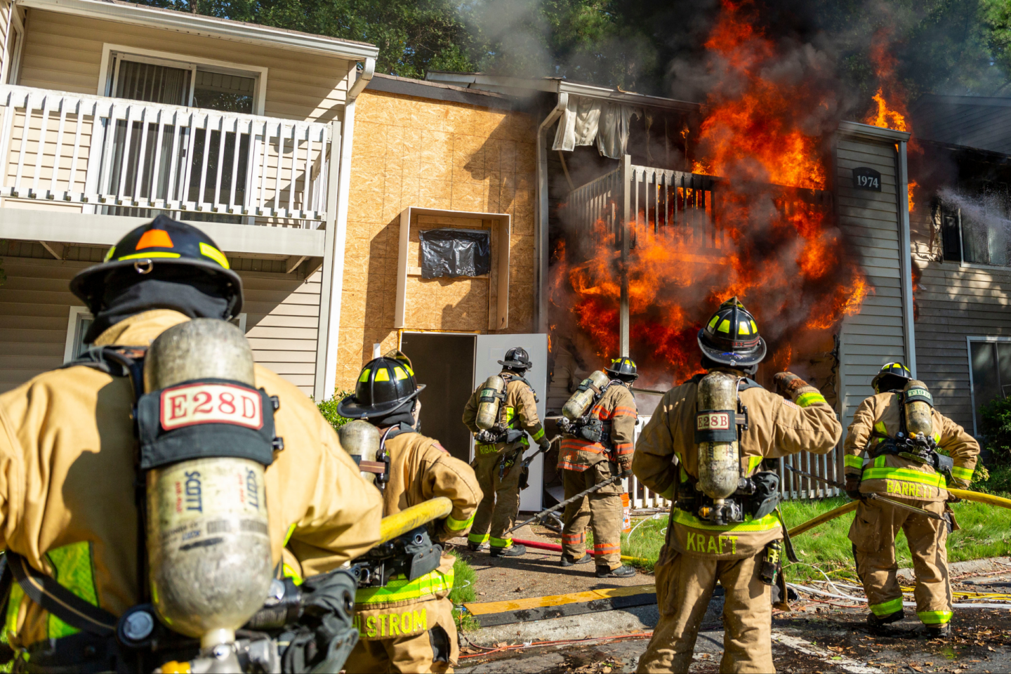 Six firefighters seen from behind assess a fire in a multi-story apartment building while advancing hoselines from the ground. Smoke and flames are visible from multiple units as crews coordinate fire attack operations, reflecting concepts taught in the Evidence-Based Structural Firefighting Course.