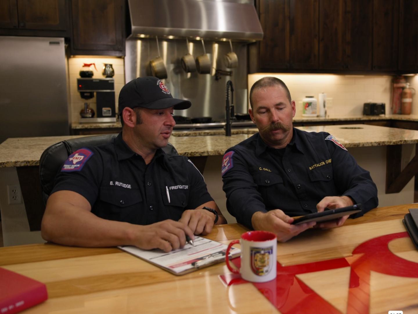 Two firefighters sit at a kitchen table in a fire station, reviewing firefighting training together on a tablet held by one firefighter while both look at the screen.